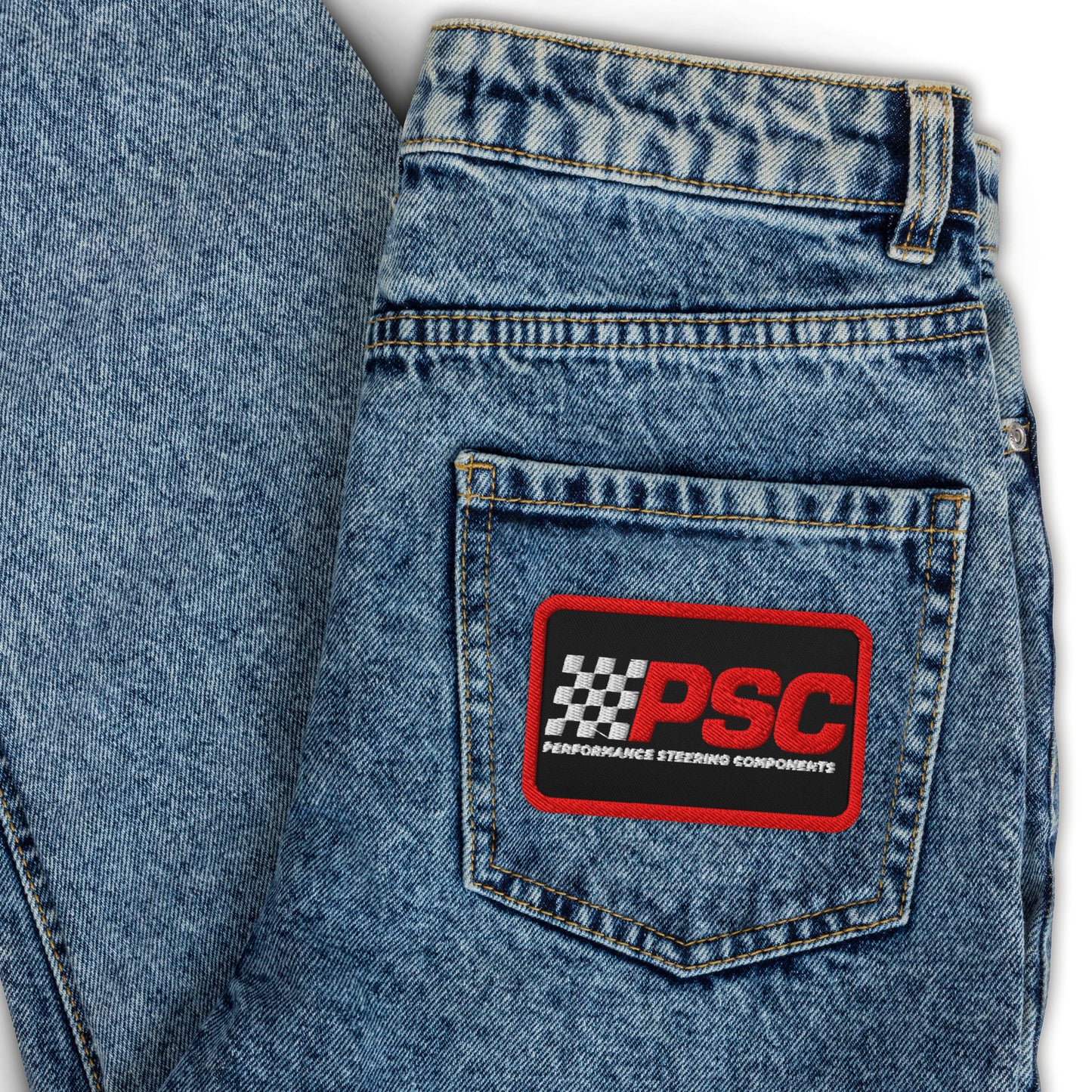 Image of jeans folded over showing a black (with red border) PSC logo in red and white with black background on the jeans pocket. The background behind the jeans is white.
