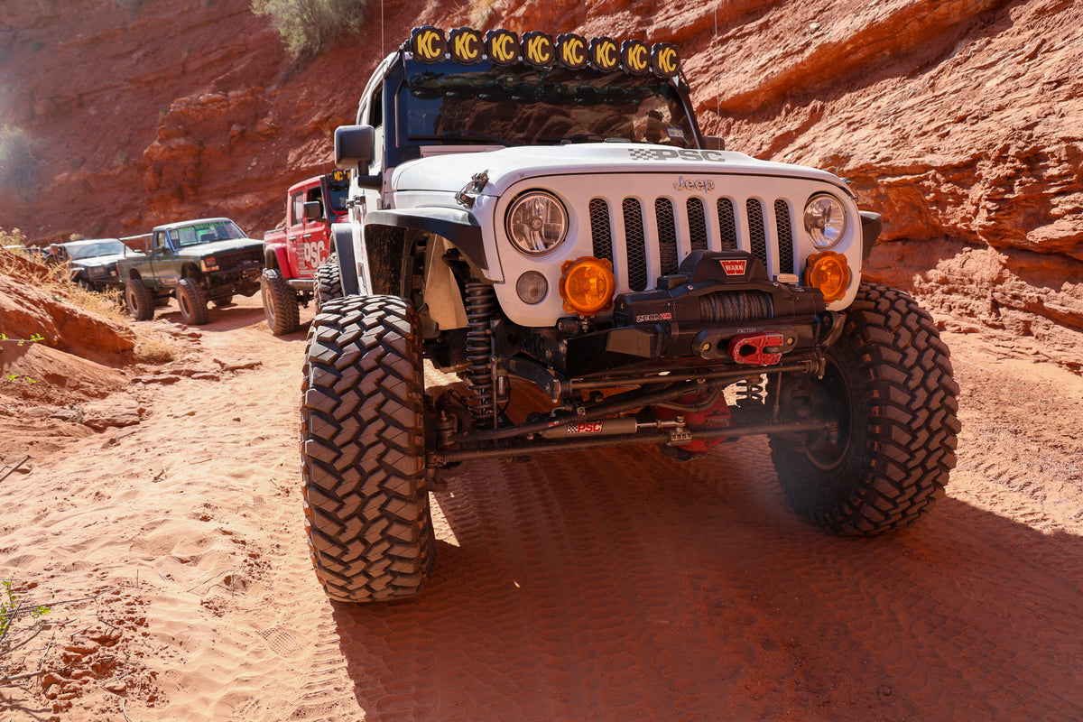 White Jeep Wrangler with PSC branding crawling through a Moab trail, showing oversized off-road tires, long-arm suspension and steering components, with additional Jeeps and trail rigs following behind.