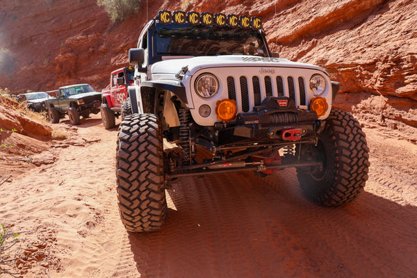 White Jeep Wrangler with PSC branding crawling through a Moab trail, showing oversized off-road tires, long-arm suspension and steering components, with additional Jeeps and trail rigs following behind.
