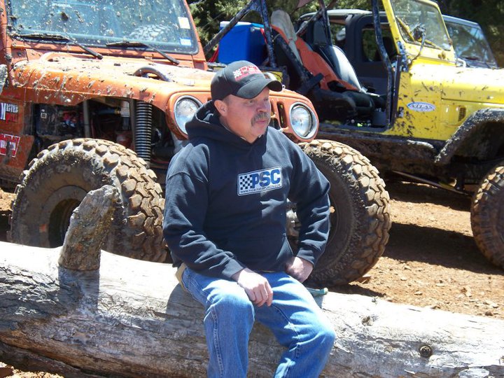 Shot of Tom Allen sitting on a log with a row of jeeps behind him. He is wearing a black hoodie with a blue PSC logo on it and a black hat with a ed and white PSC logo. 