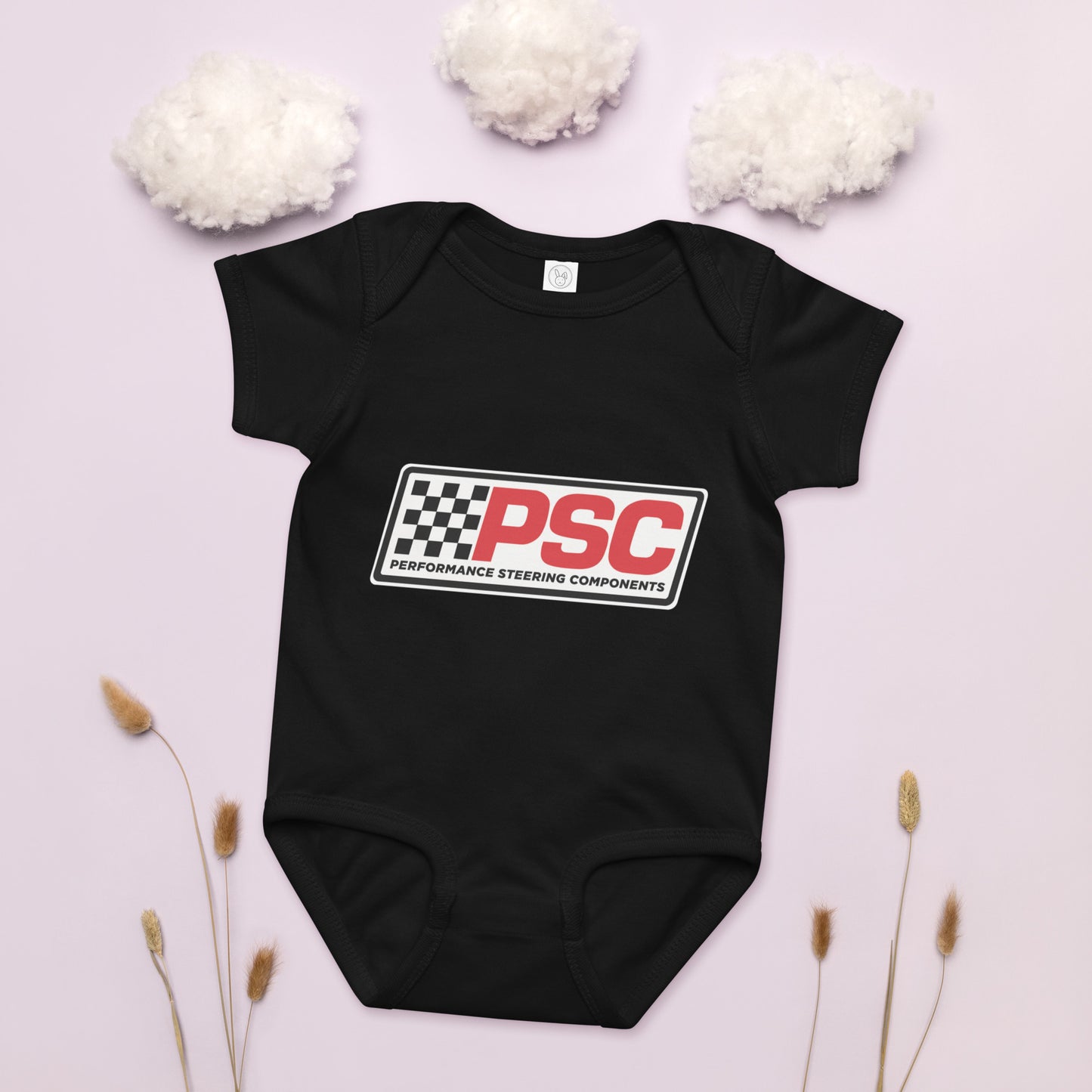 PSC logo black baby bodysuit, jersey material product mockup with a pink background showing cotton as clouds and some plants on the bottom for decoration. 