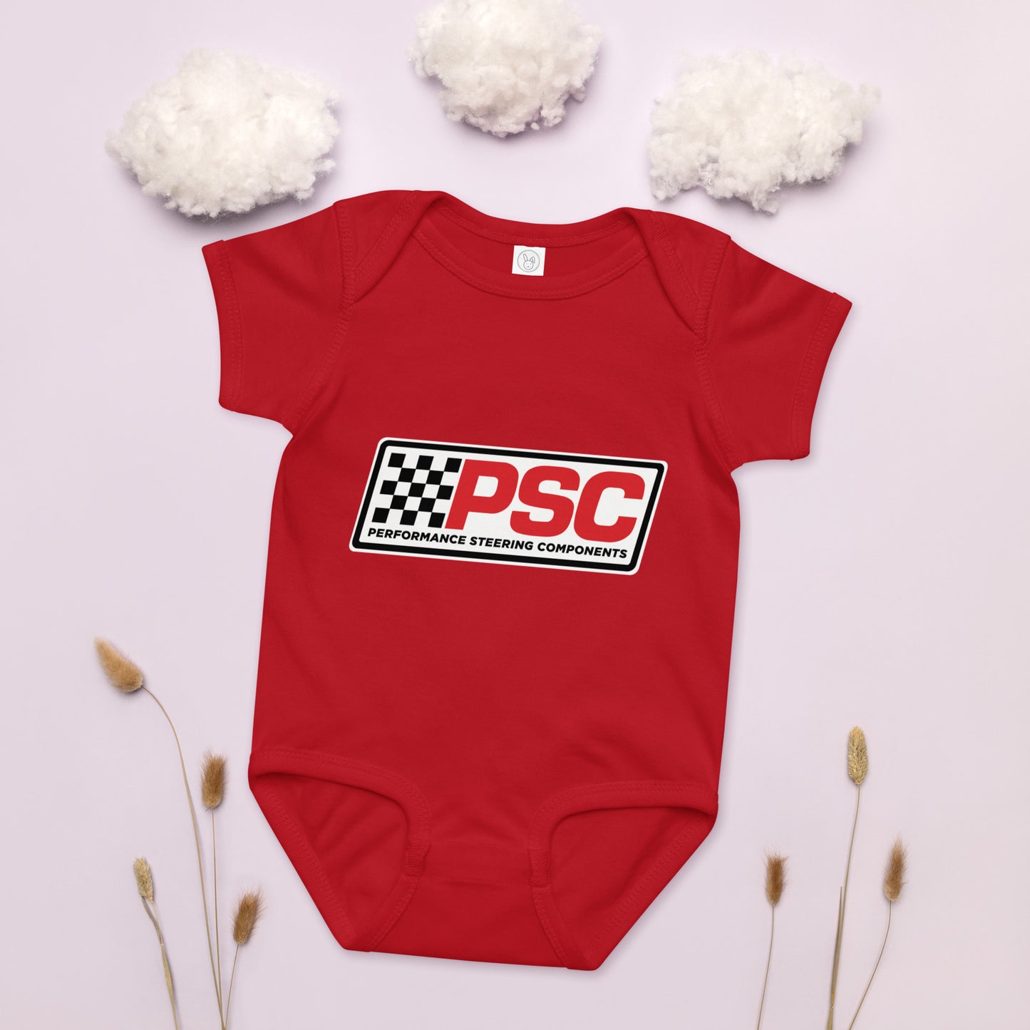 Product PSC logo red baby bodysuit, jersey material product mockup with a pink background showing cotton as clouds and some plants on the bottom for decoration. 
