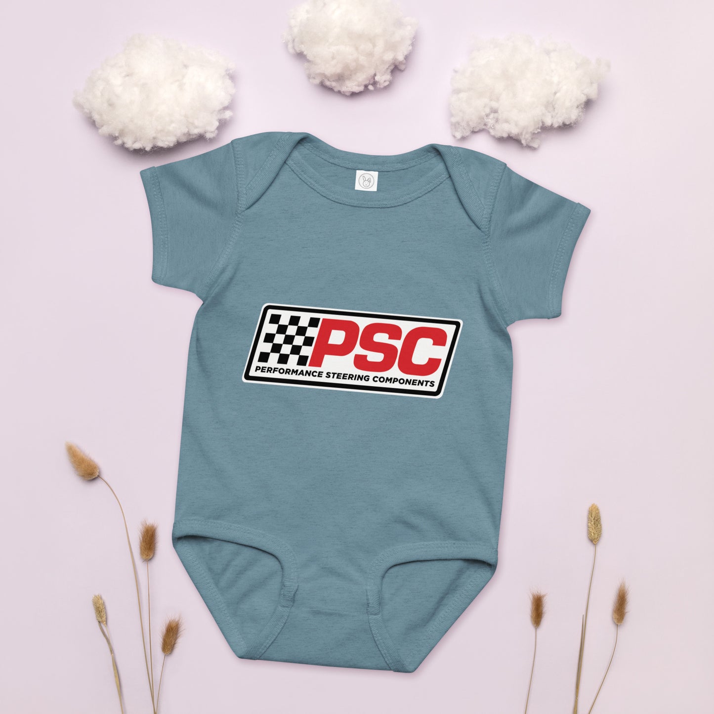 PSC logo vintage indigo baby bodysuit, jersey material product mockup with a pink background showing cotton as clouds and some plants on the bottom for decoration. 
