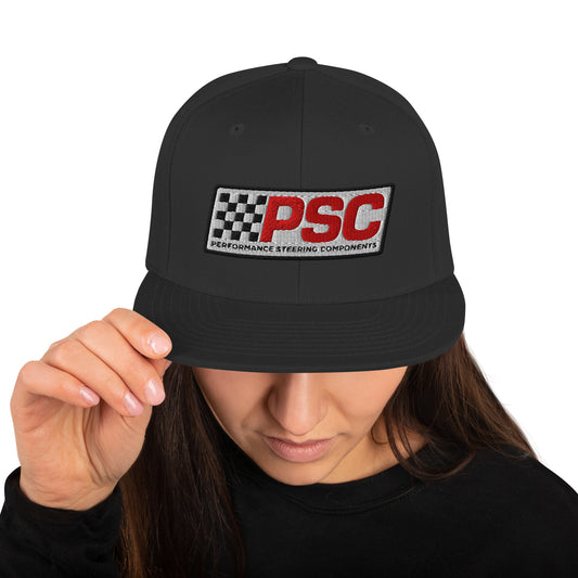 Product mockup image of the back of a classic snapback cap in a black color with black snap clasps, accents and bill. The model is wearing a black t-shirt and background behind her is white. The PSC logo with "PSC" in red, the flag and words "Performance Steering Components" are in back with a white background and black border are embroidered on the center of the cap.