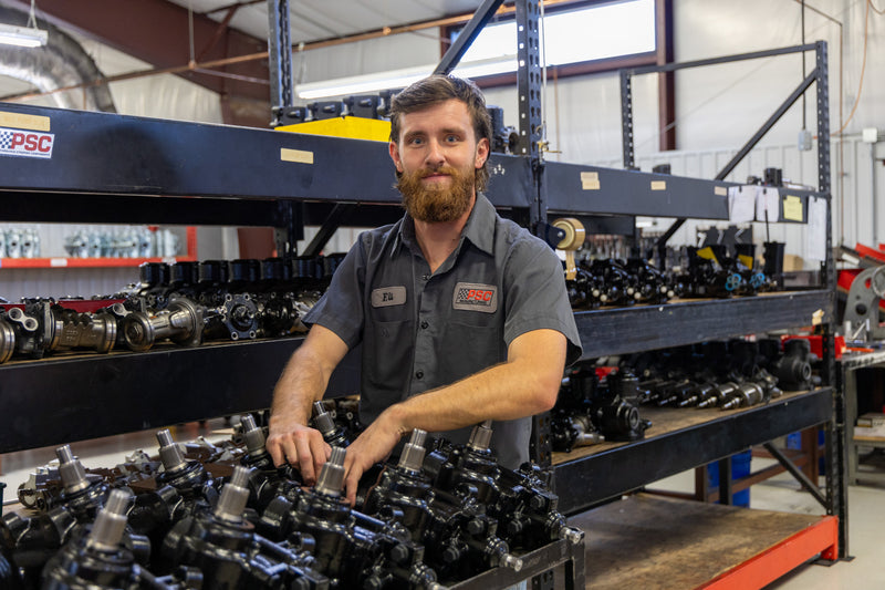 A PSC technician named Eli organizes and inspects a large rack of steering gearboxes inside the PSC manufacturing facility, surrounded by assembled components and workstations. The image showcases PSC’s precision-built steering gears, quality control process, and commitment to reliable off-road, Jeep, truck, and performance power steering solutions.