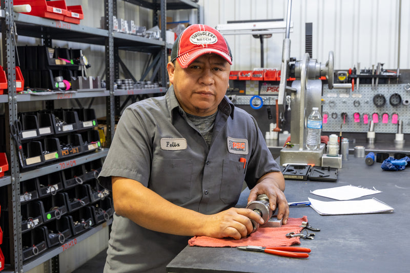 PSC technician named Felix works on a steering component inside the PSC manufacturing shop, inspecting parts on a workbench surrounded by tools, hydraulic steering components and organized inventory bins. The image highlights PSC’s precision craftsmanship, quality control, and American-made power steering solutions for off-road, rock crawling, Jeep, truck and performance applications.