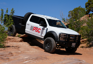 White Ford Super Duty truck with PSC branding driving over slickrock terrain, suspension flexed while off-roading under a clear blue sky.