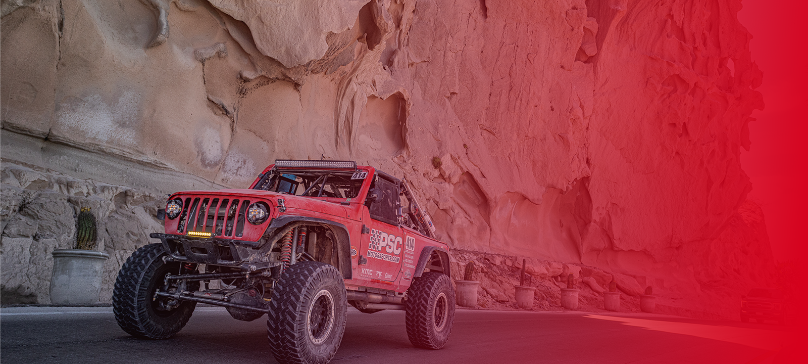 Picture of a red PSC branded off-road Jeep driving on a road along a desert canyon wall during the 2025 Norra 1000 raced, showcasing heavy-duty steering updates built for extreme terrain. The photo includes a red-to-transparent gradient from the right side of the screen. 