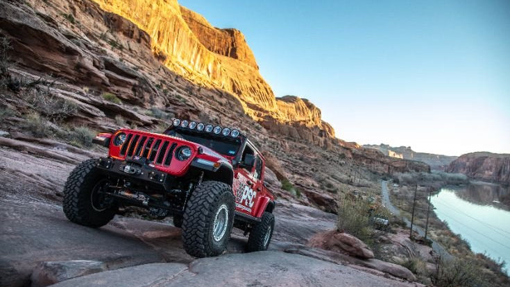 Red Jeep Wrangler equipped with large off-road tires and roof-mounted lights driving across sloped slickrock near a river canyon.