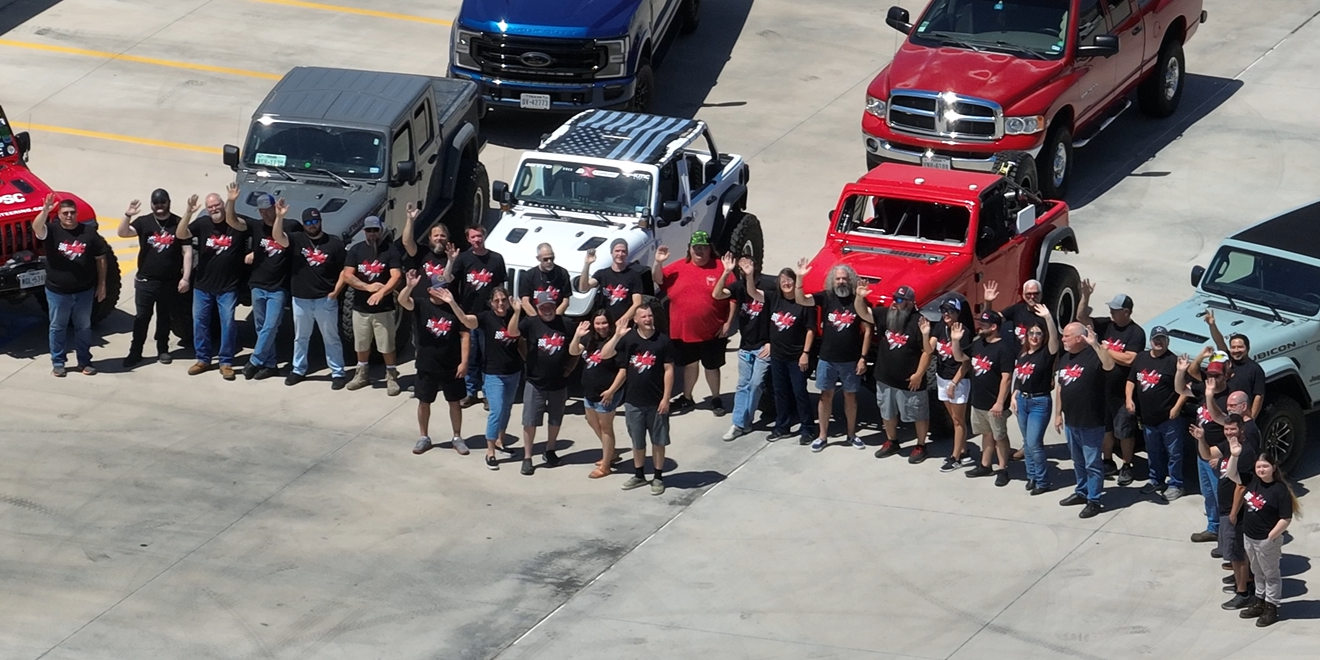 Zoomed in overhead drone shot showing PSC staff waving at the drone. You can see various jeeps packed in a semi-circle between buildings one and two. The image is slightly pixelated and blurry. 