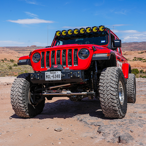 Red JT Rubicon with PSC branding is parked on a hill on a trail in Moab, Utah during EJS 2025. There is a clear shot of the PSC steering cylinder and steering components on the front-end with a blue sky and Moab hills and mountains in the background. 