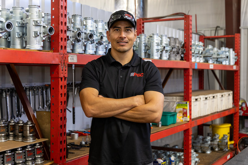 PSC Manufacturing lead, Tony, stands with arms crossed in front of organized shelves of power steering components inside the PSC facility, highlighting precision-built parts, quality workmanship, and PSC’s commitment to reliable off-road, Jeep, truck, and performance steering solutions.