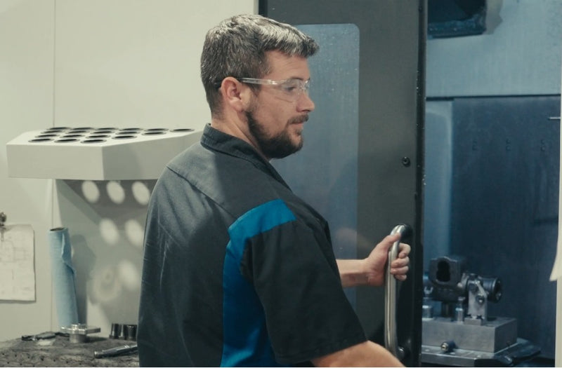 Photo of a guy in a dickies style work shirt, featuring a blue stripe running vertically, closing a door on a CNC machine, you can see the shell of a steering gear in the machine. 