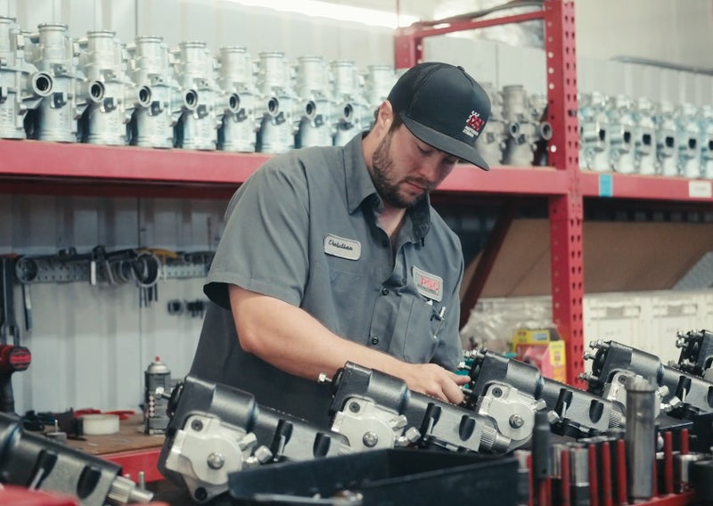 Shot of a guy wearing a black hat, grey workshirt with the name tag showing "Christian" and the PSC logo, he's screwing in bolts by hand on a line of black steering gear. There is a slight haze on the image. 