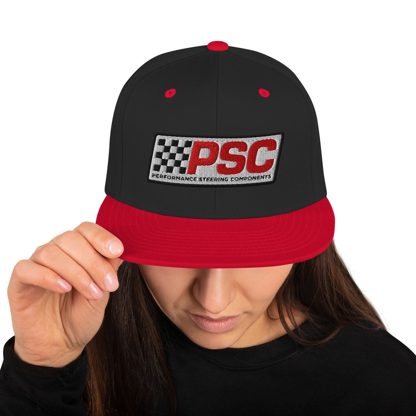 Product mockup image of the front of a classic snapback cap in a black color with red snap clasps, accents and bill. The PSC logo with "PSC" in red, the flag and words "Performance Steering Components" are in back with a white background and black border are embroidered on the center of the cap. The model is wearing a black t-shirt and background behind her is white.
