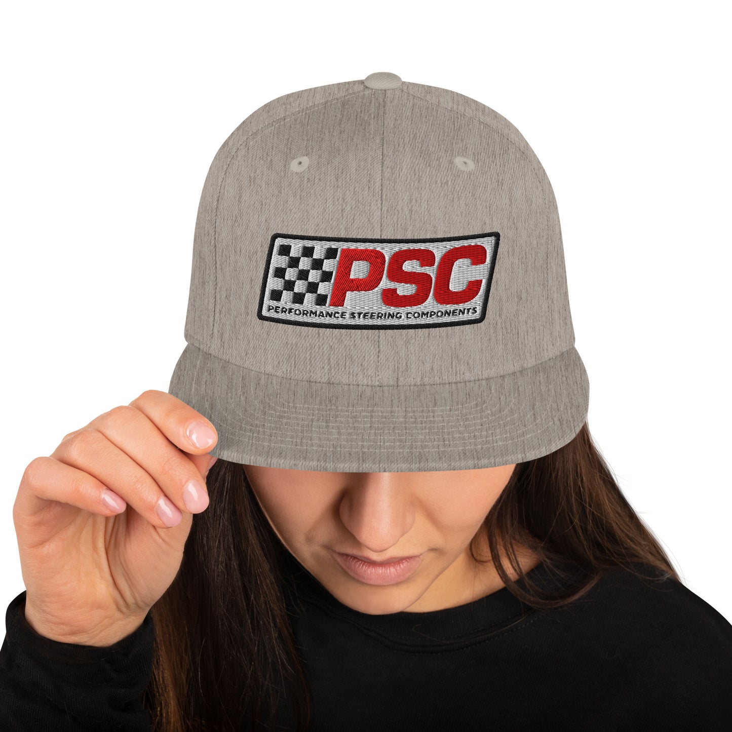 Product mockup image of the front of a classic snapback cap in a heather grey color with heather grey snap clasps and bill. The PSC logo with "PSC" in red, the flag and words "Performance Steering Components" are in back with a white background and black border are embroidered on the center of the cap. The model is wearing a black t-shirt and background behind her is white.
