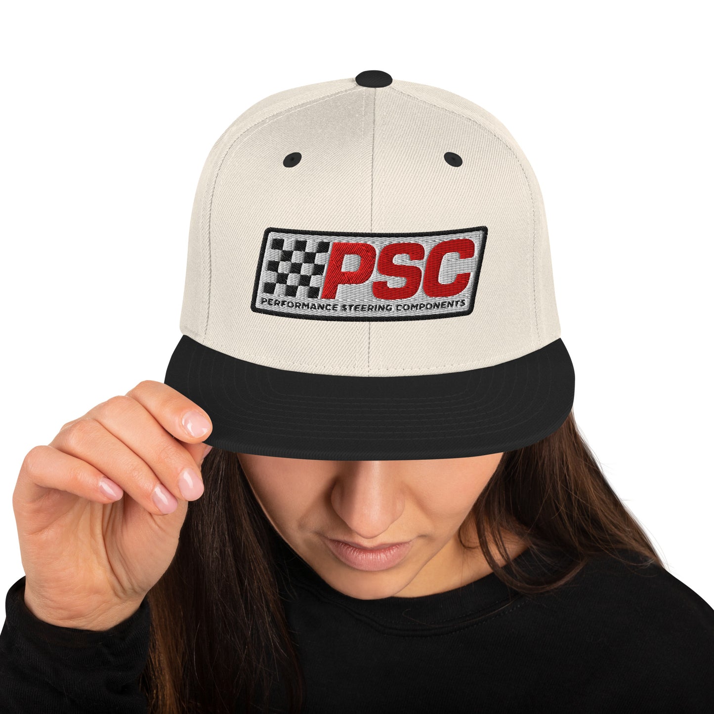 Product mockup image of the front of a classic snapback cap in a neutral tan color with black snap clasps and black bill. The PSC logo with "PSC" in red, the flag and words "Performance Steering Components" are in back with a white background and black border are embroidered on the center of the cap. The model is wearing a black t-shirt and background behind her is white.
