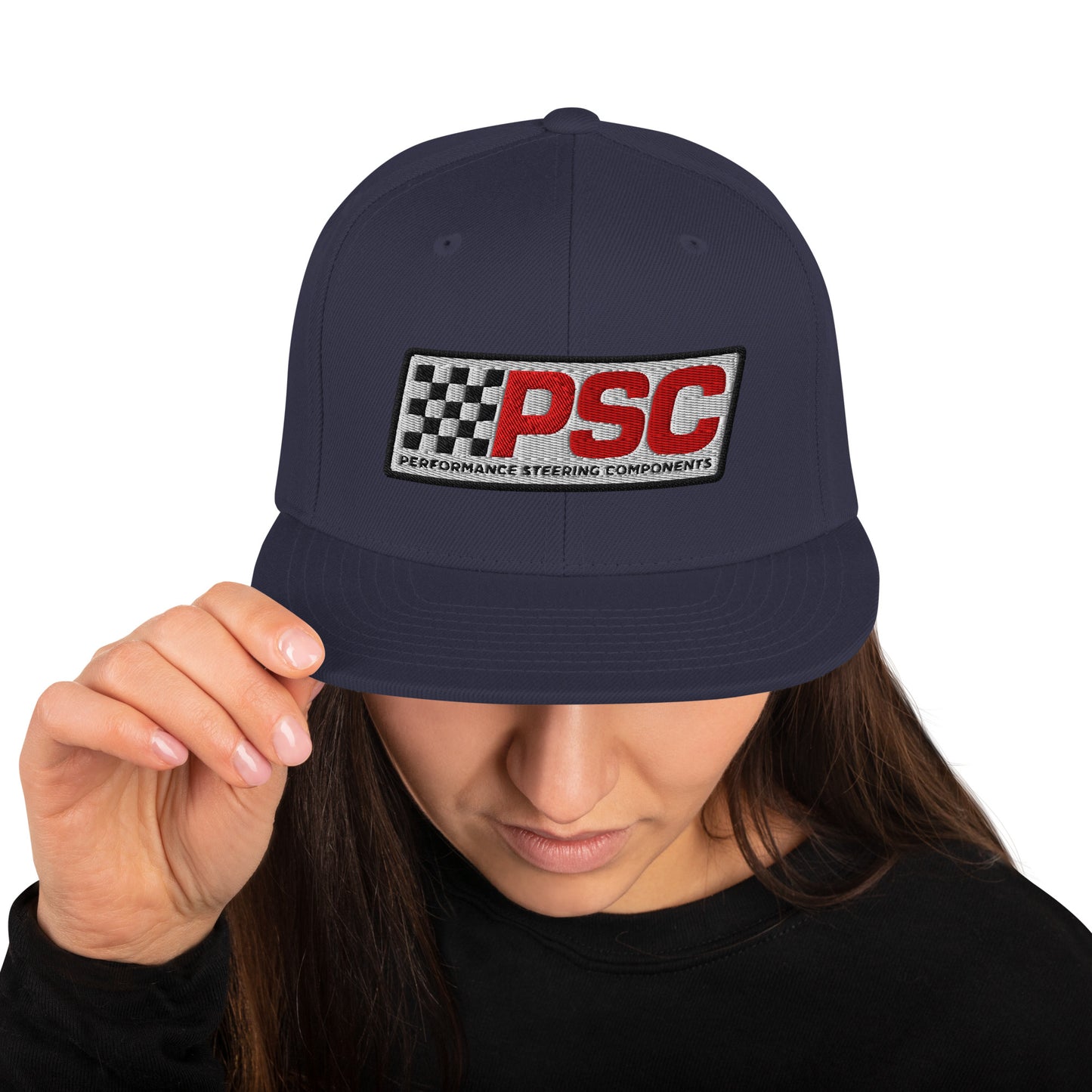 Product mockup image of the back of a classic snapback cap in a navy blue color with navy blue snap clasps and black bill.  The model is wearing a black t-shirt and background behind her is white. The PSC logo with "PSC" in red, the flag and words "Performance Steering Components" are in back with a white background and black border are embroidered on the center of the cap.