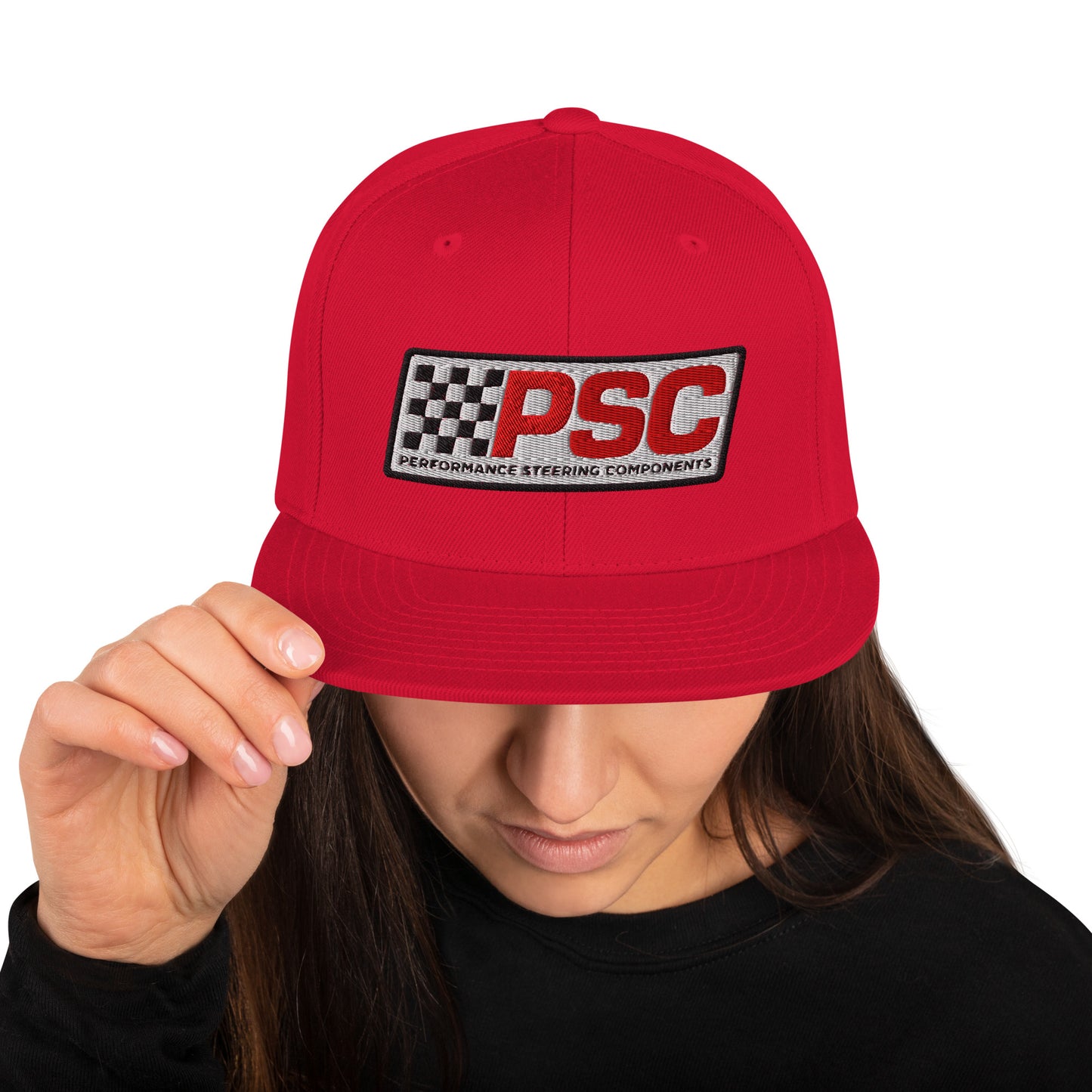 Product mockup image of the front of a classic snapback cap in a red color with red snap clasps, accents and bill.  The PSC logo with "PSC" in red, the flag and words "Performance Steering Components" are in back with a white background and black border are embroidered on the center of the cap. The model is wearing a black t-shirt and background behind her is white.