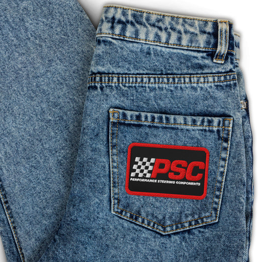 Image of jeans folded over showing a black (with red border) PSC logo in red and white with black background on the jeans pocket. The background behind the jeans is white. 