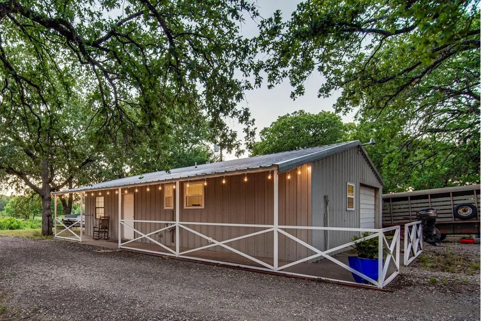 Shot of a longer building with a shop door on the right side and farmer trailer parked outside. There are lights hanging from the porch and tree all around. 
