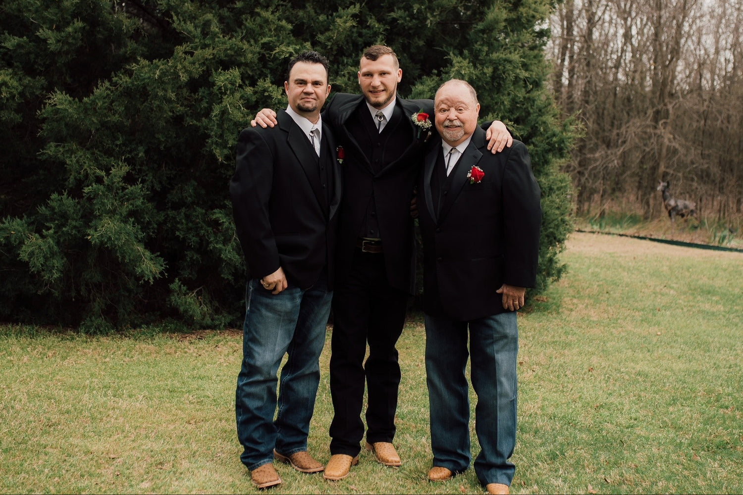 Photo of Jeff, Josh and Tom Allen all wearing black suite jackets with red rose boutonnieres, embracing and smiling in front of a large evergreen tree  with more trees in the background. 