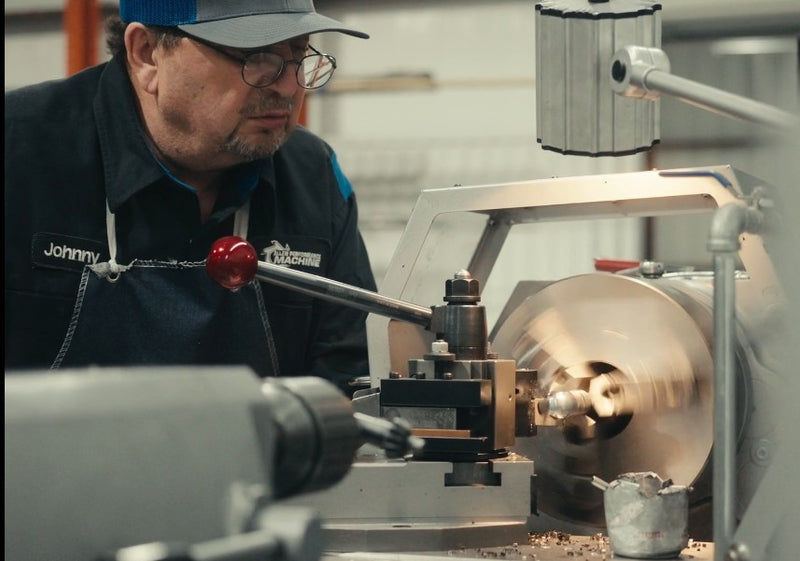 Shot of a guy wearing a blue and grey hat, black workshirt with the name tag showing "Johnny". Johnny is running a piece of machine that is on and shaves off a little amount of metal off the piece on it. There is a slight haze on the image. 