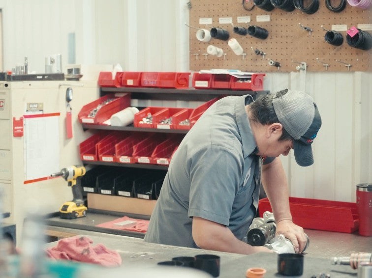 Shot of a guy wearing a black and grey hat, grey workshirt. He is working on a piece of steering gear that is sort of out of sight on a bench. There is a slight haze on the image. 