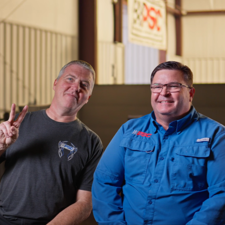 Photo of two men on screen in PSC research and development shop with a PSC banner hanging the the upper background. The one on the left has grey hair and blue eyes and is in a heathered grey PSC t-shirt holding up a peace sign. The one on the right has darker hair, is smiling and wearing glasses and a vibrant blue fishing shirt with "PSC embroidered" on the right pocket. 