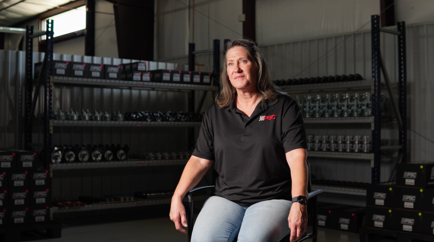 Kim Allen, President of PSC Motorsports is in a chair smiling at the videographer off screen. She's wearing a black polo with the white and red PSC logo on the left pocket area. In the background are shelves with PSC steering components and gear. 