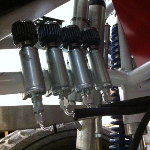 Close-up of four metallic hydraulic reservoirs with black caps mounted on a vehicle frame, alongside a blue coil spring and metal tubing.