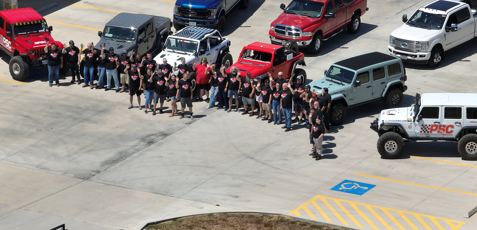 Overhead drone shot showing PSC staff waving at the drone. You can see various jeeps packed in a semi-circle between buildings one and two. The image is slightly pixelated and blurry. 
