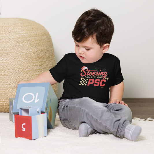Young child sitting on the floor wearing a black PSC ‘Steering All the Way’ toddler T-shirt, with holiday-themed graphics and checkered design visible on the front. Behind the words there are translucent snowflakes. There is a Santa hat on the "S" in steering and Christmas bells hanging off the "S" and "g" in steering with a "2025 under all the graphics.