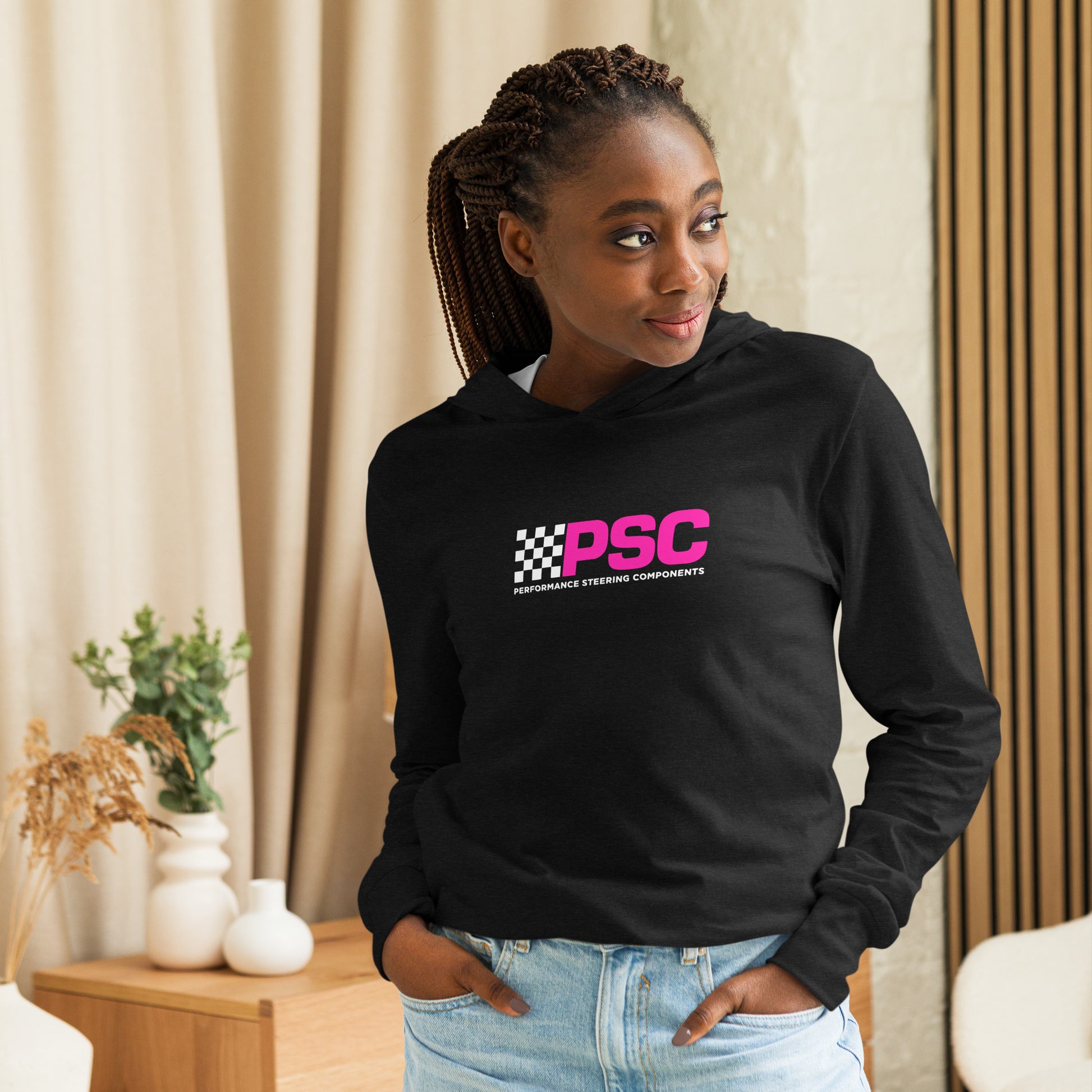Product mark up of a model wearing a black unisex long-sleeve t-shirt with hood attached featuring PSC logo centered on the front. The “PSC” are in bold and bright pink with a white flag to the left with the words “Performance Steering. She is smiling on looking to the right of the frame. There is decorations for a dresser and vases in the background behind her. 