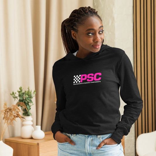 Product mark up of a model wearing a black unisex long-sleeve t-shirt with hood attached featuring PSC logo centered on the front. The “PSC” are in bold and bright pink with a white flag to the left with the words “Performance Steering. She is smiling on looking to the right of the frame. There is decorations for a dresser and vases in the background behind her. 